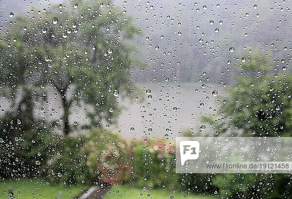 Wolkenbruchartiger Regen  durch die Fensterscheibe betrachtet  Hintergrund Garten und Weiher in Unschärfe