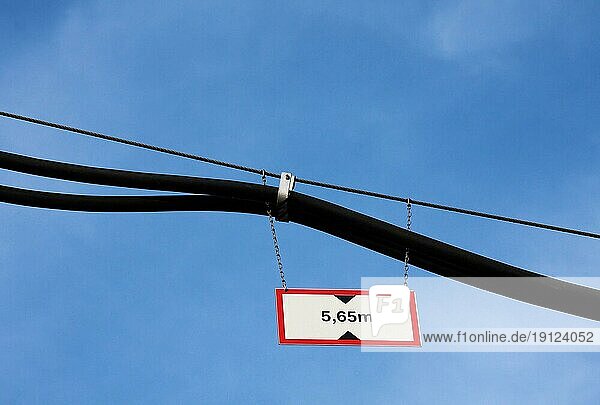 Rot-weiss-schwarzes Verkehrsschild an einer Metallkette an Kabeln hängend mit der Aufschrift 5  65 m als Hinweis zur Durchfahrt