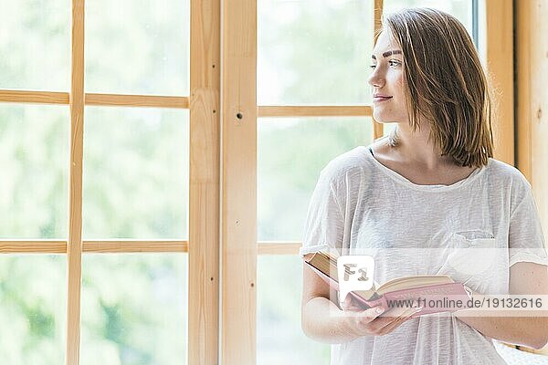 Hübsche junge Frau steht vor dem Fenster und hält ein Buch
