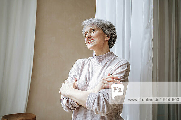Smiling mature woman with arms crossed standing by curtain at home