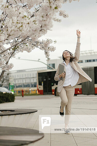 Cheerful businesswoman jumping by cherry blossom flower tree