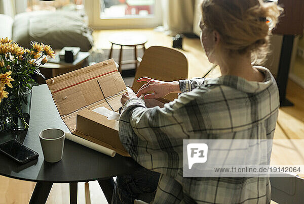 Young woman unpacking cardboard box on table at home