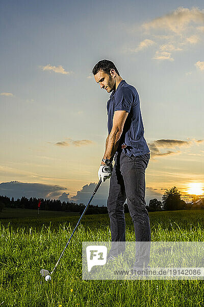 Young man preparing to take golf shot on grass at dusk