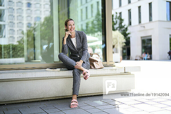 Smiling businesswoman with hand on chin sitting on bench near glass wall