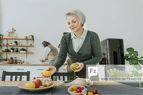 Smiling woman arranging fruits on dining table in kitchen at home
