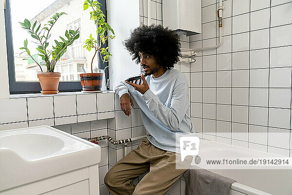 Man talking on speaker phone sitting on bathtub in bathroom at home