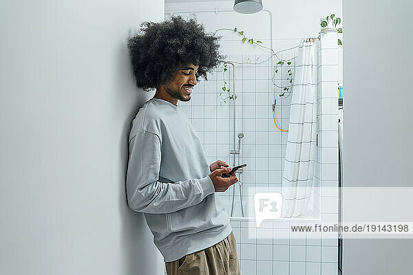Smiling man using smart phone leaning on wall in bathroom at home