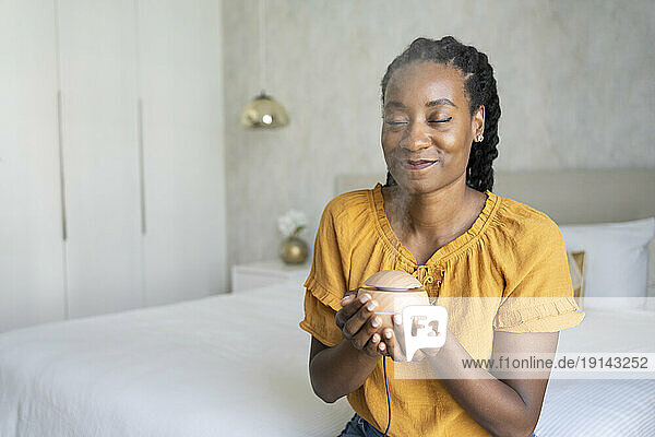 Smiling woman smelling aroma from air diffuser sitting on bed at home