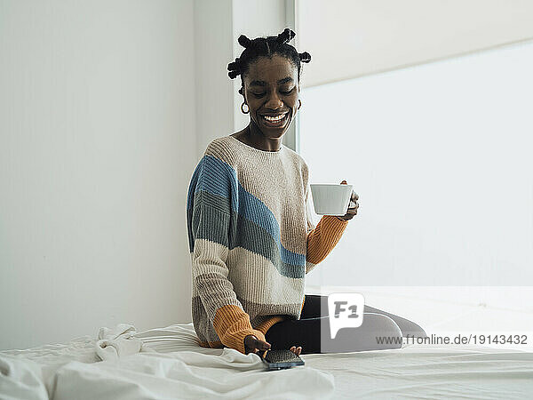 Smiling woman holding coffee cup and using smartphone on bed