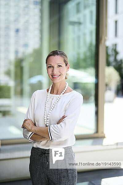 Smiling businesswoman standing with arms crossed in front of glass wall