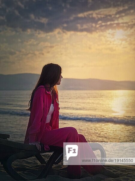 Sorglose junge Frau ästhetisches Porträt sitzen entspannt auf der Sonnenliege am Strand mit Blick auf den Sonnenaufgang über dem Meer. Wunderschöne Szene der Morgendämmerung am Meer. Sommerurlaub  Reisen und Entspannung Konzept