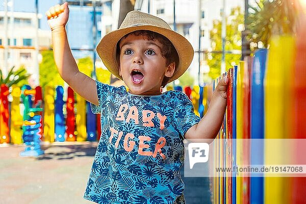 Porträt eines glücklichen kleinen Jungen mit Hut  der auf einem Spielplatz spielt und die Hand hebt  um etwas zu feiern