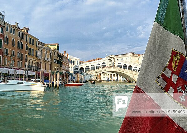 Schöner Blick auf die italienische Seeflagge und die Rialtobrücke oder Ponte Rialto am Canal Grande in Venedig  Italien. Gondel Service Station auf der linken Seite. UNESCO Weltkulturerbe Stadt. Tageslicht  Sonnenschein  weiche Wolken. Selektiver Fokus