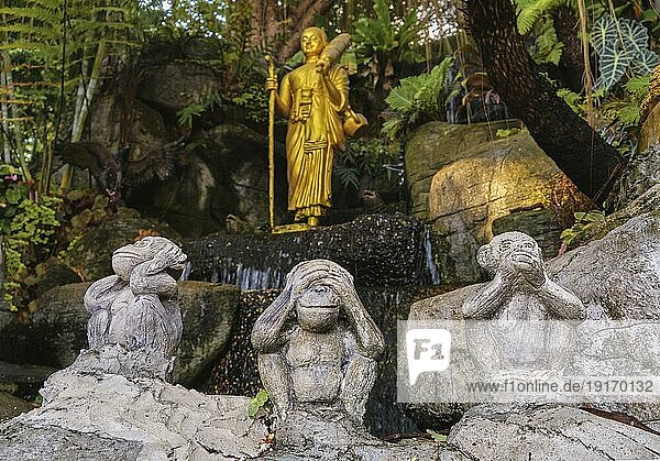 Buddhismus Symbole: vergoldete Buddhastatue als wandelnder Mönch mit Naga Wanderstab und Schirm sowie drei weise Affenstatuen im Vordergrund. Religion  Vielfalt  Inklusivität  Gebet  Anbetung