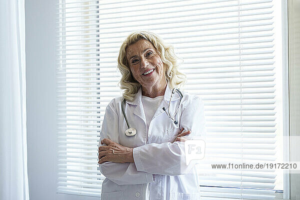 Happy senior doctor standing with arms crossed in front of window
