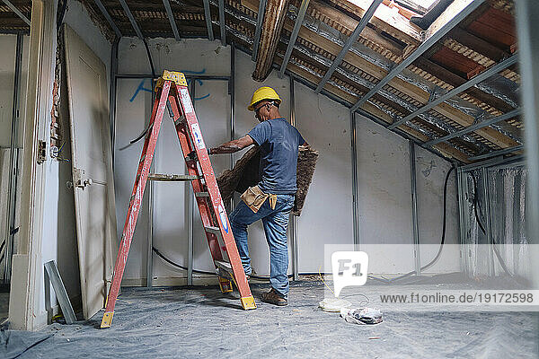 Mature construction worker climbing on ladder with insulation at site