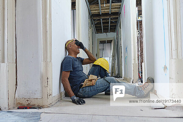 Happy mature construction worker covering eyes with hand sitting in corridor