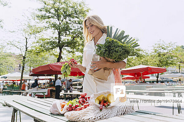 Happy woman with various vegetables standing at table in market