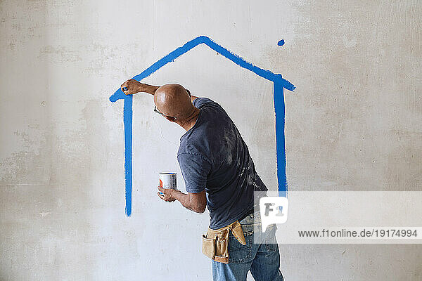 Bald construction worker drawing house on wall with paintbrush at site