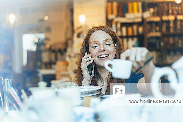 Smiling redhead woman talking on smart phone and shopping for crockery at store