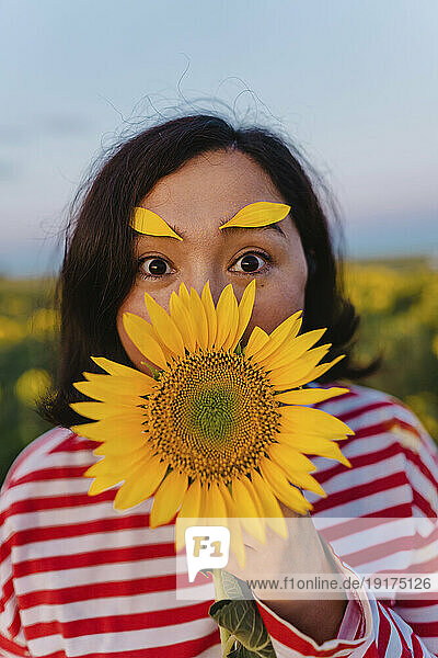 Mature woman with petals on eyebrows covering mouth with sunflower in field