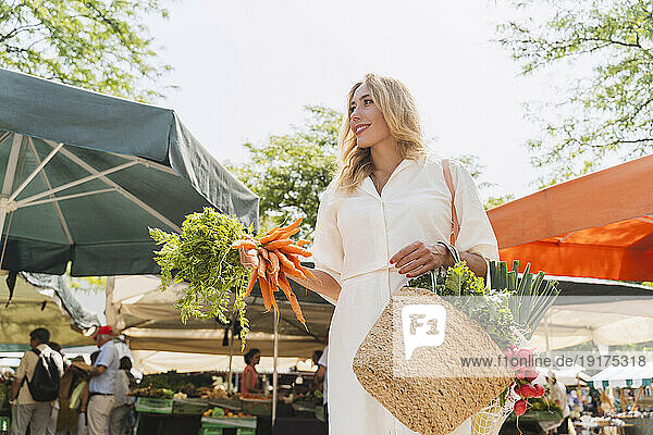 Smiling woman holding bunch of carrots and bag of groceries in market