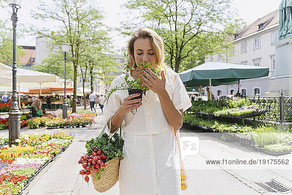 Smiling blond woman smelling mint plant and holding groceries on sunny day