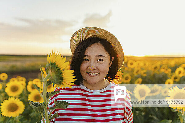 Smiling woman with hat holding sunflower in field at sunset