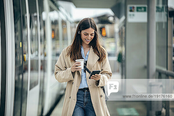 Happy woman walking with coffee cup and using smart phone at tram station