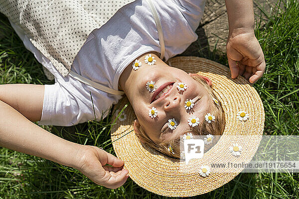 Happy boy with daisies over face relaxing on grass