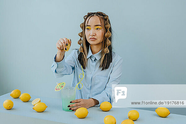 Thoughtful woman squeezing lemon in cocktail against blue background