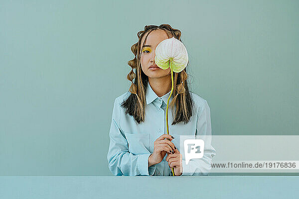 Young woman covering eye with flower against blue background