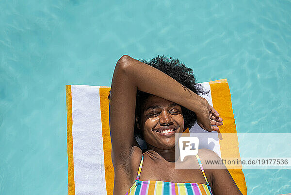 Smiling young woman lying on stripped towel with eyes closed in pool