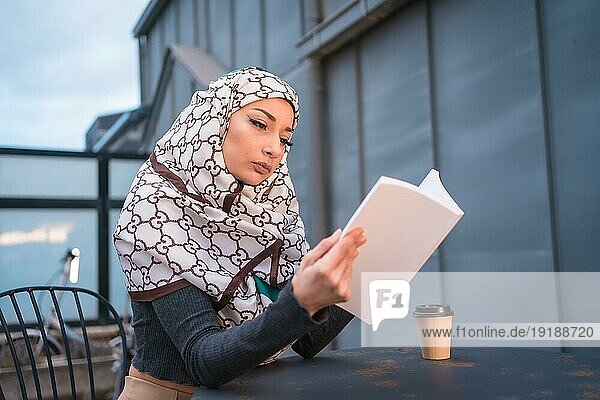 Arabisches Mädchen mit weißem Schleier am Computer auf der Terrasse eines Coffeeshops  das mit viel Spaß ein schönes Buch liest