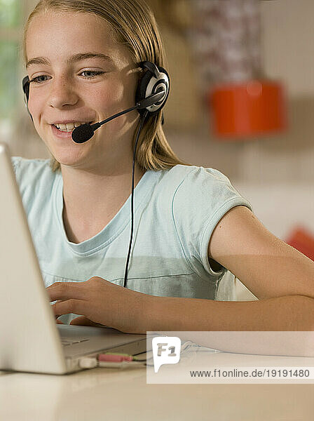 Smiling young girl wearing earphones and microphone sitting at desk typing on a laptop computer