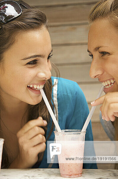 Two smiling teenaged girls drinking milk-shake with a straw from the same glass
