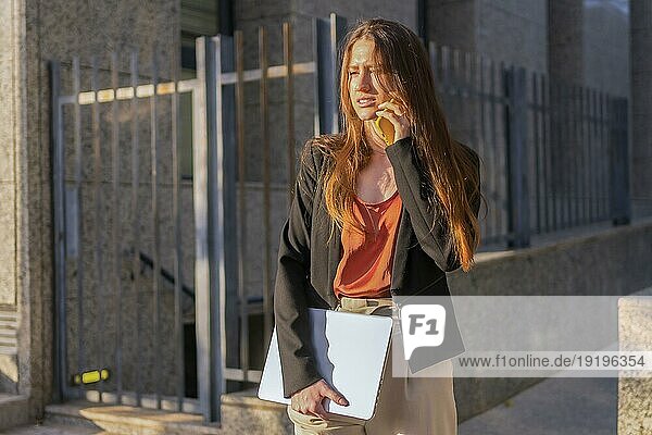 Lässige Geschäftsfrau im Gespräch mit dem Handy im Freien  während sie einen Laptop trägt