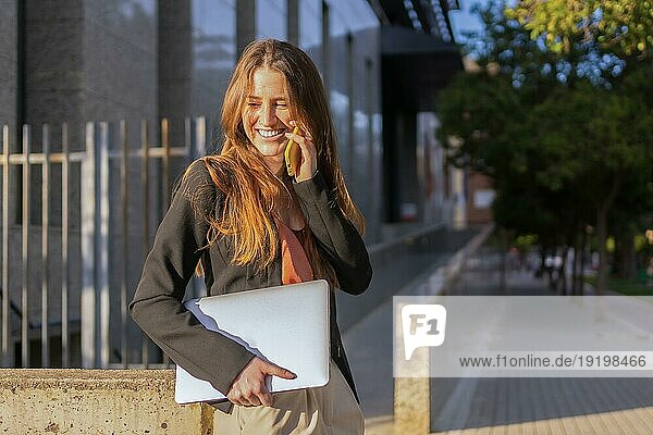 Porträt einer lächelnden und lässigen Geschäftsfrau  die mit dem Mobiltelefon spricht und einen Laptop im Freien trägt