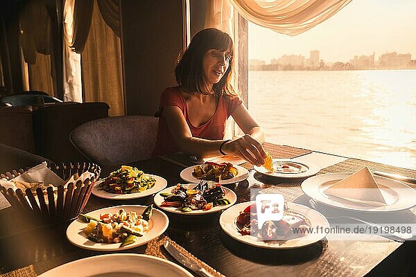 Ein junger Tourist  der auf einem Boot auf dem Nil zu Abend isst ein traditionelles ägyptisches Essen im Licht des Sonnenuntergangs. Afrika