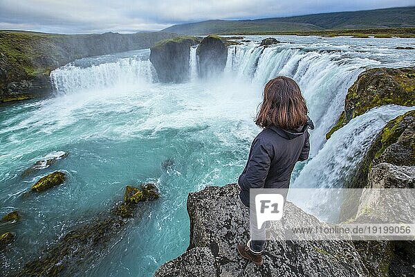 Ein junger Tourist betrachtet den Godafoss Wasserfall von oben. Island