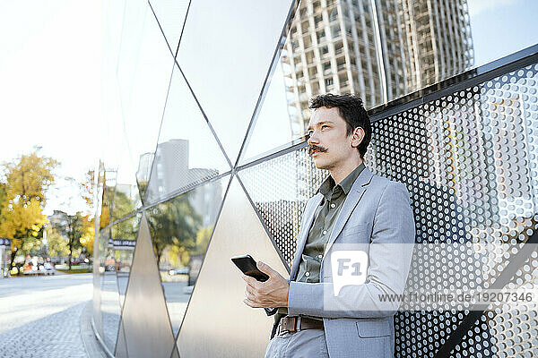 Thoughtful businessman holding smart phone in front of modern glass building