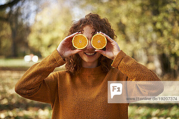 Smiling redhead woman holding orange slices in front of face at autumn park