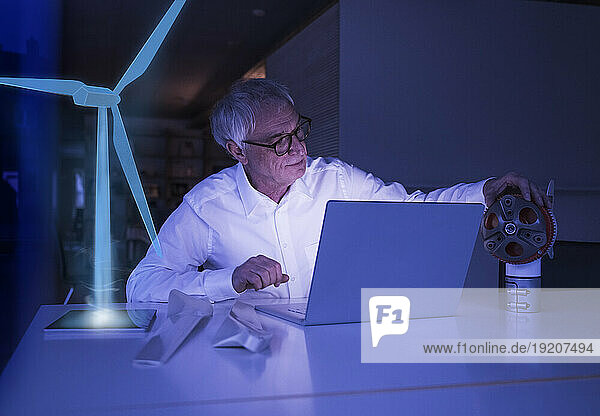 Businessman with laptop examining machine part at desk
