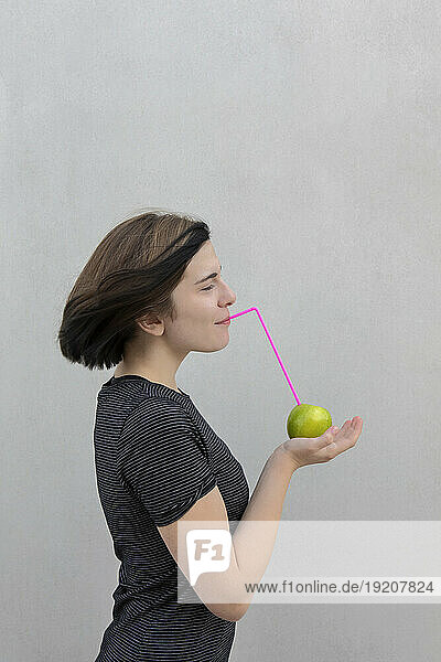 Teenage girl holding Granny Smith Apple with straw against gray background