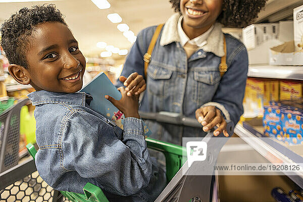 Smiling boy sitting in shopping cart with mother at supermarket