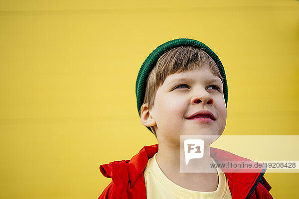 Thoughtful boy wearing red jacket and knit hat in front of yellow wall