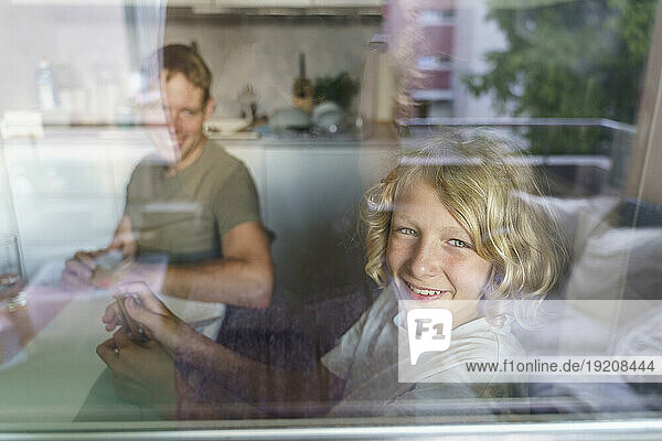 Smiling father and son looking through window from kitchen