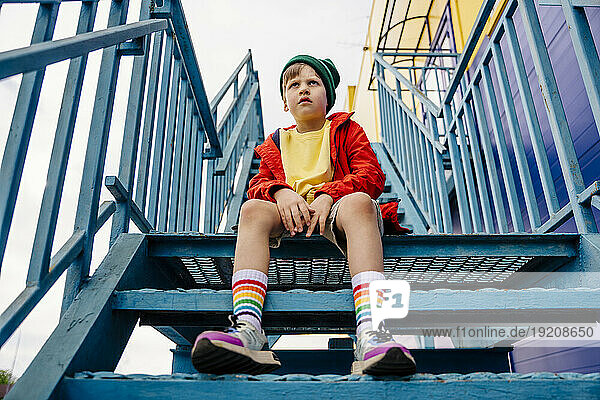 Thoughtful boy in red jacket sitting on metallic staircase