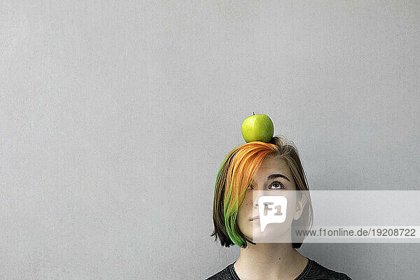 Teenage girl with dyed hair balancing apple on head against gray background