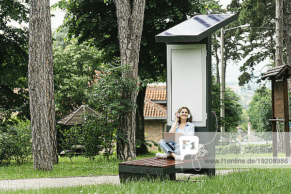 Woman sitting on bench and listening to music near solar charging point at park